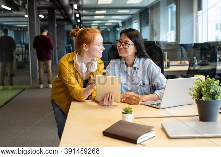 Two young intercultural female managers looking at one another with smiles while working together over presentation by workplace in office