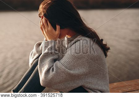 Side View Of Depressed Young Female In Warm Sweater Covering Face With Hands While Sitting On Bench 