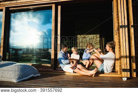 Family With Small Children Sitting On Patio Of Wooden Cabin, Holiday In Nature Concept.