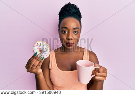 Young african american woman eating doughnut and drinking coffee relaxed with serious expression on face. simple and natural looking at the camera. 