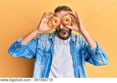 Attractive man with long hair and beard holding tasty colorful doughnuts on eyes in shock face, looking skeptical and sarcastic, surprised with open mouth 