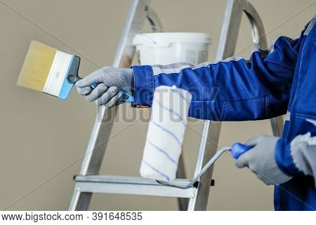 The Plaster Painter Is Ready To Paint The Wall. In The Hands Of A Roller And Brush. A Stepladder And