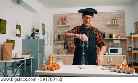 Grandfather Sprinkling On The Table Sifted Flour In Modern Kitchen. Elderly Senior Baker With Bonete