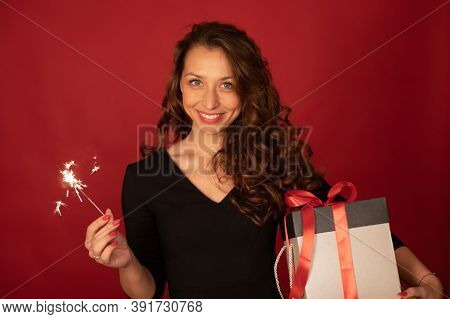 Young Charming Lady With Gift Box And Burning Sparkler Smiling Looking At Camera On Red Background. 