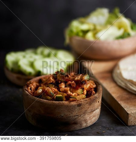 a bowl with a chicken meat and vegetables filling on a table, next to a plate with some slices of cucumber, a bowl with lettuce and some tortillas or durums to prepare burritos or wrapping sandwiches