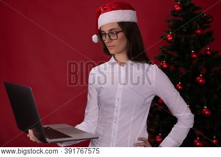 Thoughtful Brooding Brunette Girl With Long Hair In Glasses And Santa Claus Hat Uses Laptop To Plan 