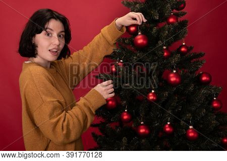Smiling Dressed Casually Brunette Girl Hanging Toy Balls On Christmas Tree On Studio Red Backdrop. T