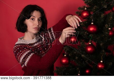 Brunette Girl Dressed In Knitted Winter Pattern Sweater Hanging Toy Balls On Christmas Tree On Studi