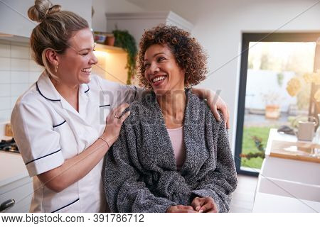 Mature Woman In Dressing Gown Talking With Female Nurse In Kitchen At Home