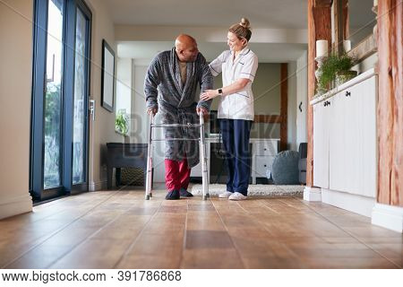 Senior Man In Dressing Gown Using Walking Frame Being Helped By Female Care Worker