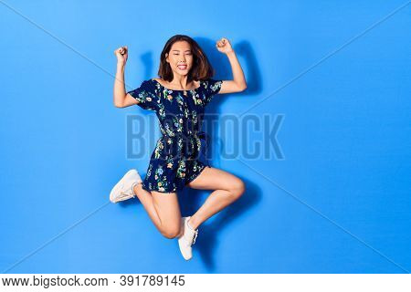 Young beautiful chinese woman wearing casual clothes smiling happy. Jumping with smile on face doing strong geture over isolated blue background