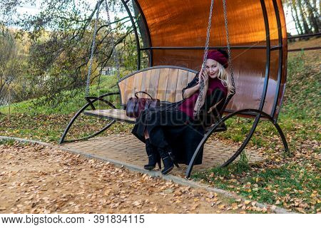 A Beautiful Girl Sits On A Garden Swing, Forged In A Burgundy Coat And Biret, With Beautiful Eyes, I