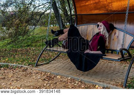 A Beautiful Girl Lies On A Garden Swing, Tossed In A Burgundy Coat And In A Biret, With A Kind Look,