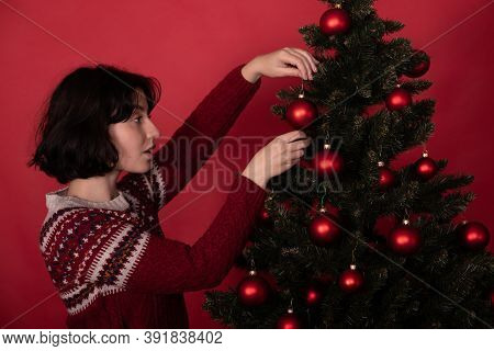 Brunette Girl Dressed In Knitted Winter Pattern Sweater Hanging Balls On Christmas Tree On Studio Re