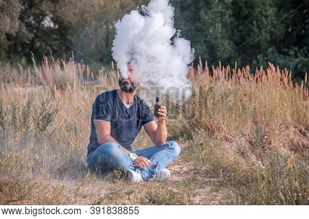Modern Man Blowing A Lot Of Smoke Using Vape Electronic Cigarette In The Forest. Man Really Likes Pr