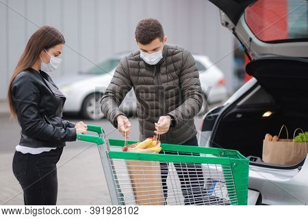 Young Stylish Couple In Protectivemask With Shopping Cart Full Of Fresh Food, Packing Products Into 