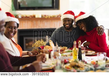 Couple wearing santa hat smiling and hugging each other while sitting and having lunch on dining table in the living room at home. christmas festivity tradition celebration concept