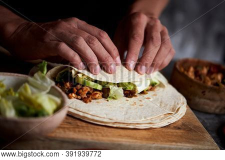 closeup of a young caucasian man preparing a durum or a burrito, with chicken meat cooked with different vegetables such as onion or red and green pepper, and fresh lettuce and raw cucumber