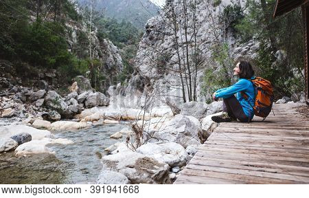 A Woman With A Backpack Sits On The Porch Of An Old Wooden House And Looks At A Mountain River. The 