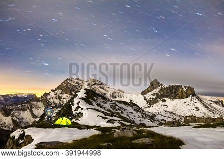 Man Standing Next To Tent Under A Starry Night Sky In Snowy Alpine Mountains. Alps, Switzerland.