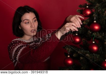 Young Sexy Woman In Sweater Decorating Xmas Tree On Red Background. New Years Eve Organized Preparat