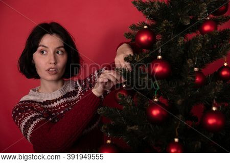 Young Pretty Woman In Sweater With Ornament Decorating Xmas Tree Looks Excited To Side, Red Backgrou
