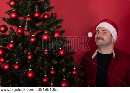 Funny Excited Man In Santa Hat Standing Near Xmas Tree Looks Satisfied At Camera On Red Background. 