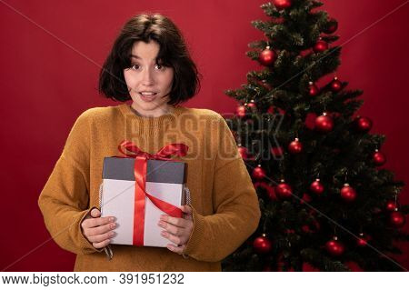 Young Pretty Woman Looks Surprised Bewildered Holding Xmas Gift Box Standing In Front Of Christmas T