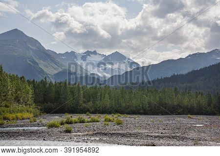 Mountains In Alaska Rise Above Trees And A Glacial Streambed On A Sunny Afternoon