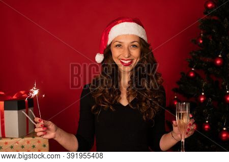 Young Smiling Woman In Santa Hat With Xmas Gifts Tree On Red Background Holding Sparkler And Champag