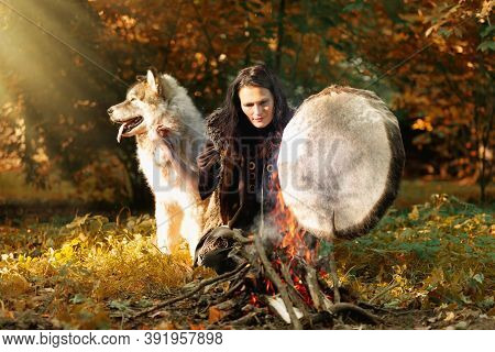 Shaman Woman Playing Her Shaman Sacred Drum In The Sunny Forest By The Fire, The Big Dog Alaskan Mal