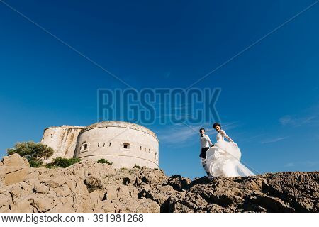 Bride And Groom Are Standing Together And Holding Hands Near The Arza Fortress On The Mamula Island