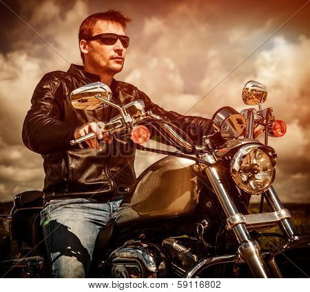 Biker man wearing a leather jacket and sunglasses sitting on his motorcycle looking at the sunset.