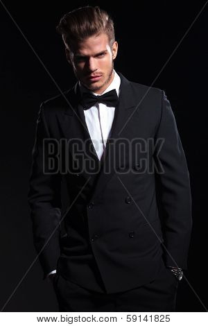 elegant young fashion man in suit holding both his hands in his pockets while looking at the camera with a frowned expression. on black background 