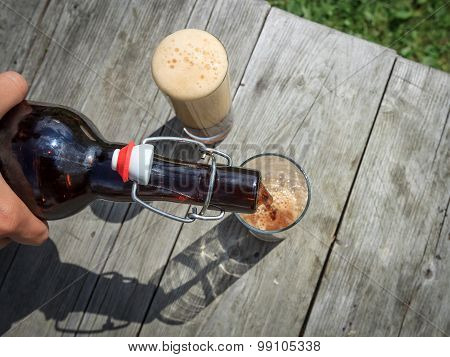 Frothy Dark Beer Pouring Into Tall Glasses From A Brown Glass Bottle In Summer Garden On Rustic Wood