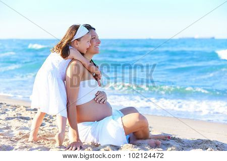 Pregnant mother and daughter on the beach together hug sitting on sand