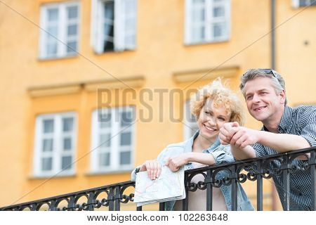 Happy middle-aged couple with map leaning on railing against building