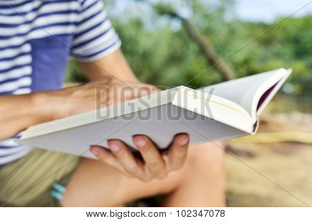 closeup of a young caucasian man reading a book outdoors in a natural landscape in a sunny day