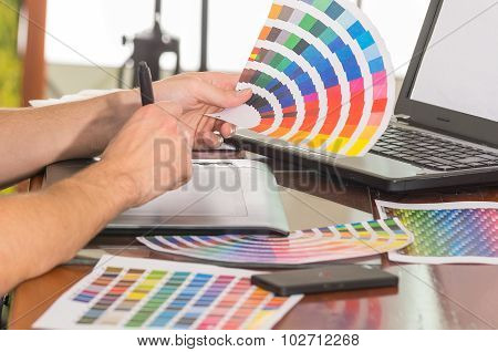 Male hands holding up palette, colormap spread out in front of laptop on working desk