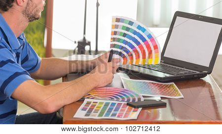 Male hands holding up palette, colormap spread out in front of laptop on working desk
