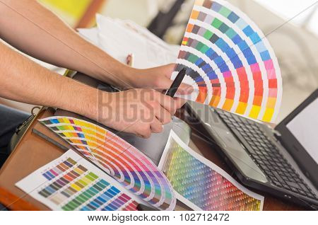 Male hands holding up palette, colormap spread out in front of laptop on working desk