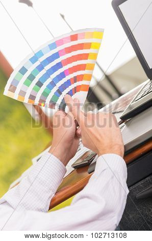 Male hands holding up palette, colormap spread out in front of laptop on working desk