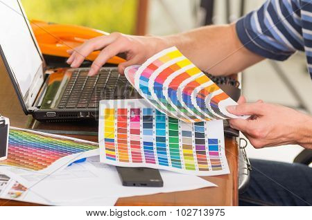 Man wearing blue white striped t-shirt sitting by work desk using phone and looking at palette, colo