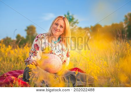 9 months pregnant woman with mehandi pattern ornament belly sitting in yellow grass and smiling on sunset lights. Waiting for baby. Pregnancy concept