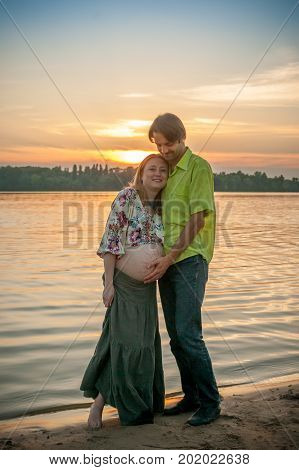 A pregnant beautiful woman with her husband on the river bank beach smiling and touching her mehandi ornament belly with love and care. Happy couple, family relax on sunset