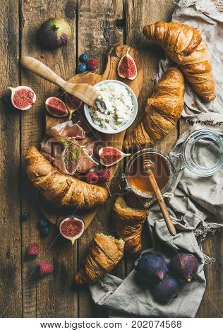 Breakfast with freshly baked croissants, ricotta cheese, figs, fresh berries, prosciutto di Parma and honey in glass jar over rustic wooden background, top view, vertical composition