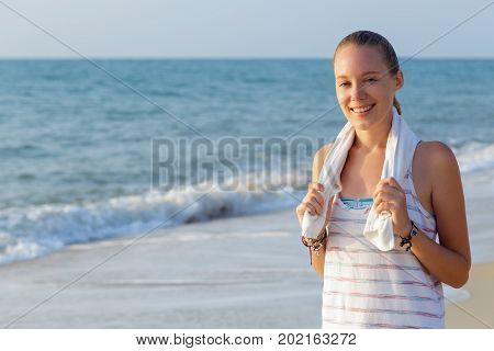 Athletic woman is standing after workout on the beach with towel around neck