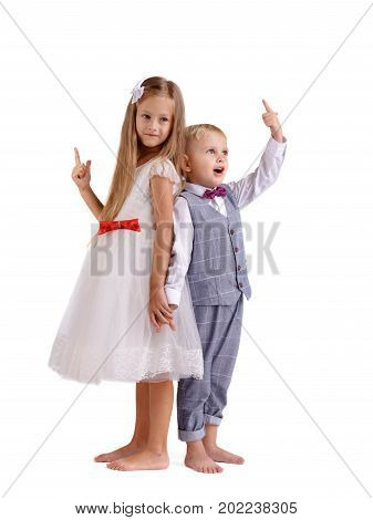 A pair of little siblings posing isolated over the white background. A charming girl in a fancy dress and a boy in a classy suit pointing at something. Friendship and happy childhood concept.