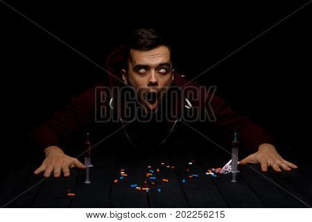 An addict sitting at the table with colorful pills and syringes on a saturated black background. An overdose of drugs. Addiction, fear, abuse and depression concept. Copy space.