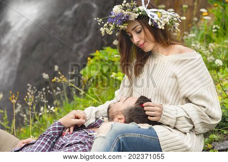 Hipster with a beard lies on the lap of his girl who wears a vinok of wildflowers. The concept of unity with nature and a young couple of people
